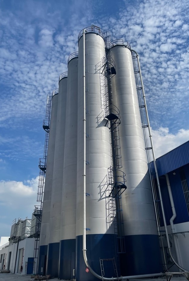 Tall cylindrical silo storage tanks outside a manufacturing facility under a partly cloudy sky.